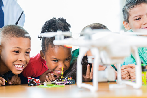 Diverse group of excited elementary students at STEM school watch as the propellers turn  on the drone they built. The group of Hispanic and African American children smile proudly as they watcch. A teacher is in the background holding the controller.